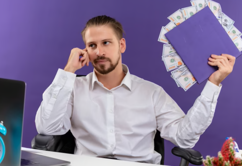 Handsome businessman in white shirt holding folder with cash looking aside with pensive expression sitting at the table in offise over purple background