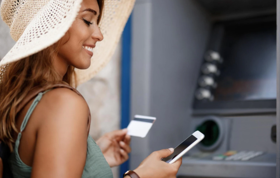 Photo smiling woman withdrawing money from cash machine while using smart phone
