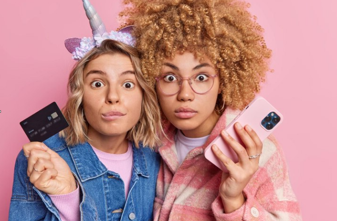 two women stare shocked at the camera holding a mobile phone and a credit card pink background 