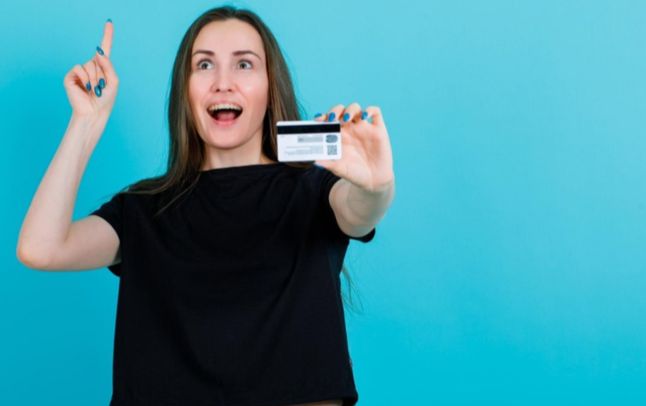 happy girl is pointing up with forefinger and showing credit card to camera on blue background
